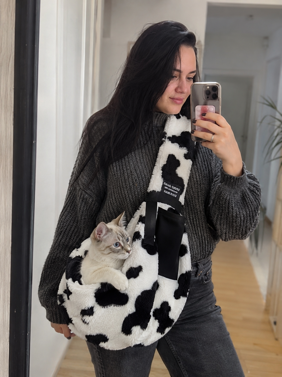Light-colored cat resting in a cow-print sling during an indoor mirror photo.