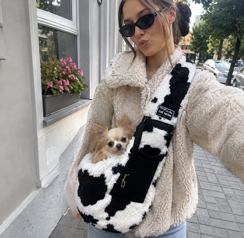 Small tan dog in a cow-print sling during an outdoor selfie.